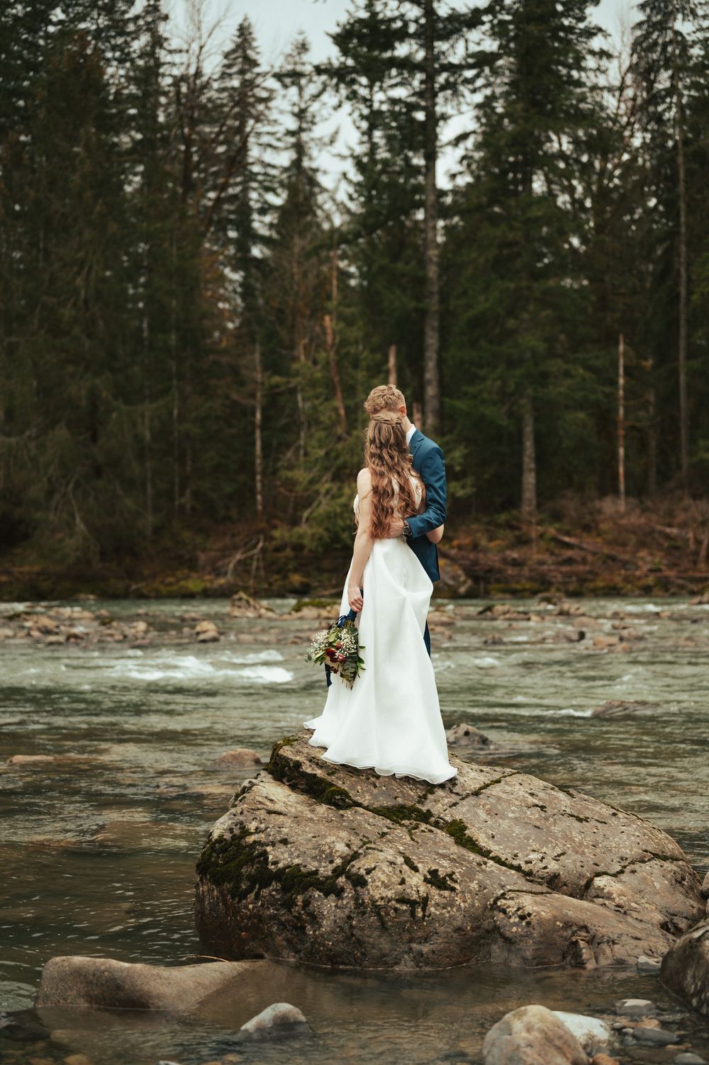 A couple stands on a rocky outcrop overlooking a rushing river surrounded by tall evergreen trees in the wilderness.