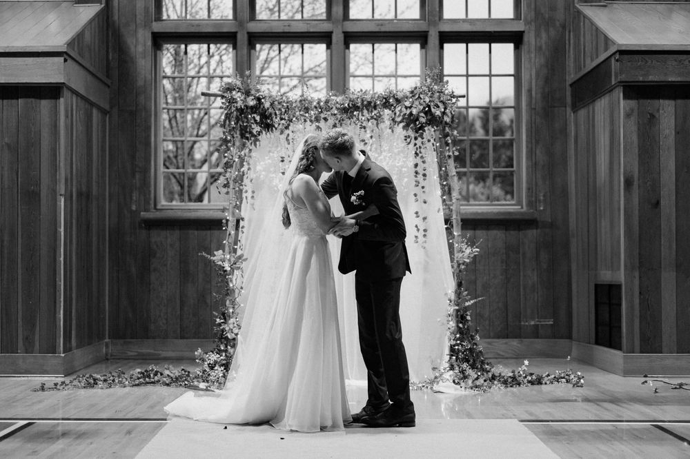 A romantic wedding ceremony silhouette under a floral arch by a large window in a rustic wooden interior.