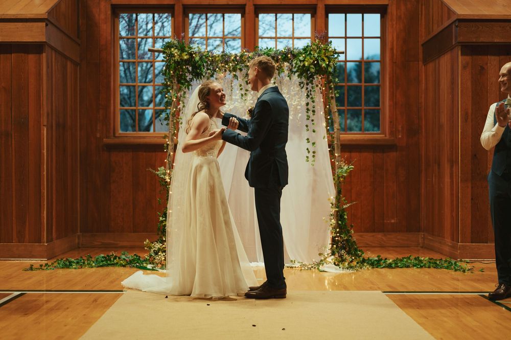 A couple shares their first dance as newlyweds beneath a floral arch in a rustic wooden venue with large windows.