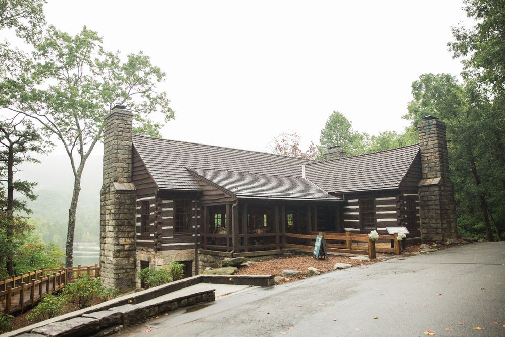 Rustic log cabin with stone chimneys and wooden porches sits on a misty hillside surrounded by green trees.