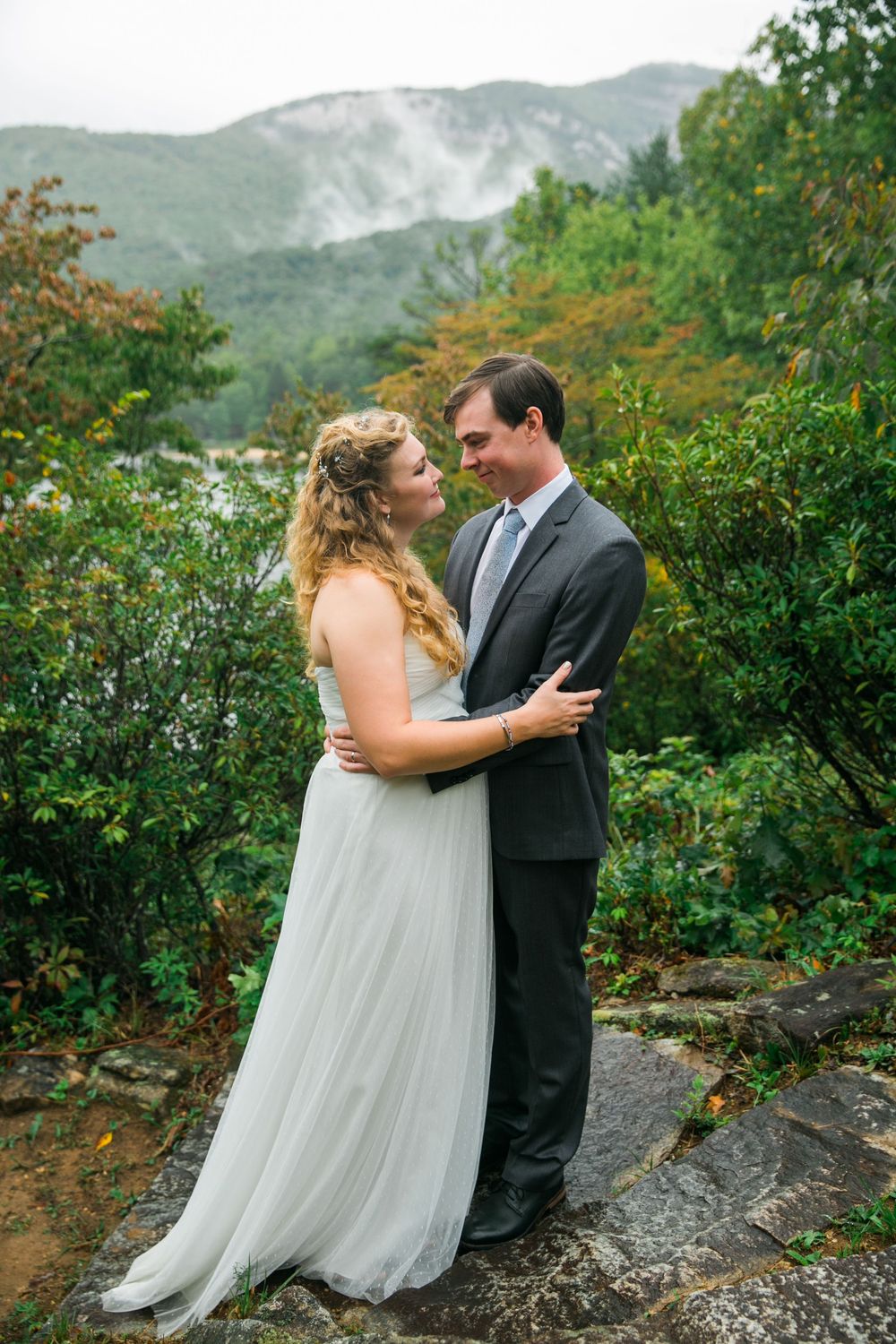 Couple embraces outdoors in scenic mountain setting with grey dress and suit against green foliage backdrop.