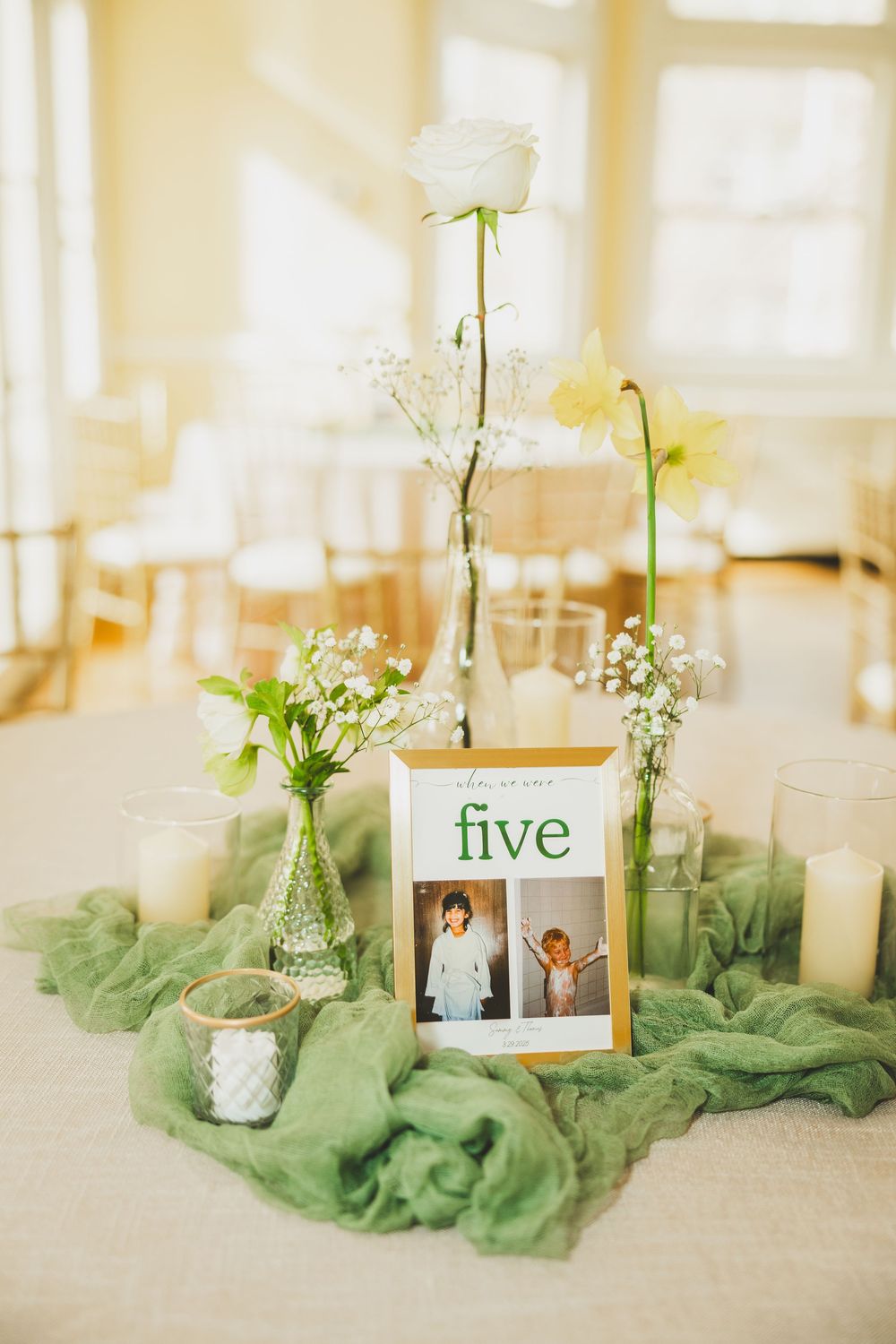 Wedding table centerpiece with white rose, baby's breath in glass vases, green tulle fabric, and table number five with photo frame.