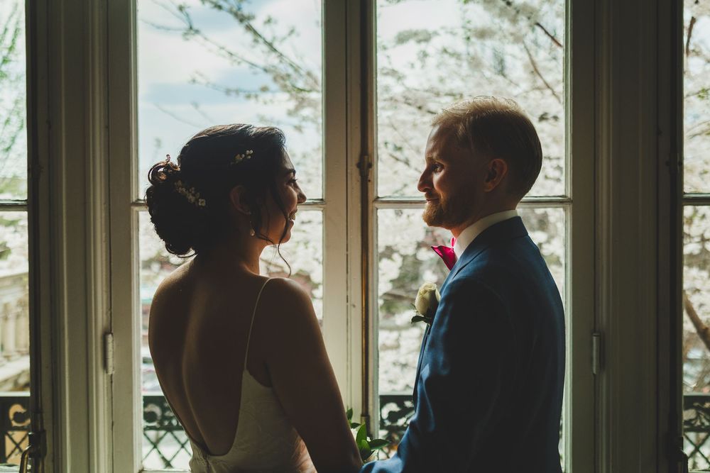 A couple stands together in front of a bright glass window while wearing formal wedding attire.