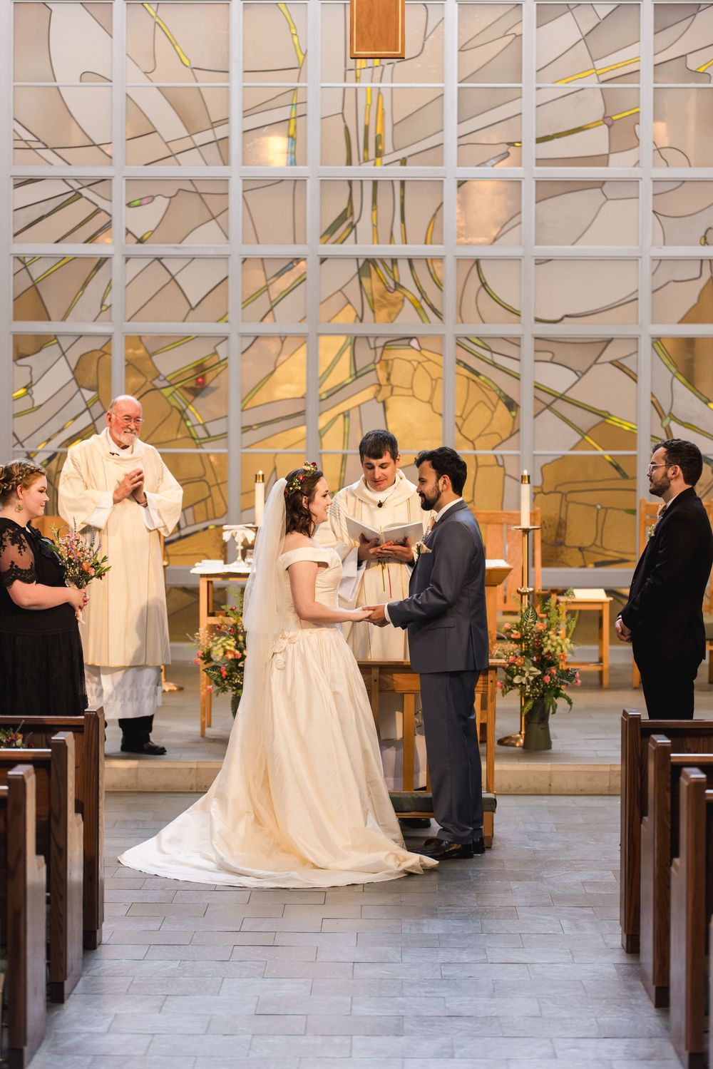 A wedding ceremony takes place in a modern church with geometric stained glass windows and floral arrangements by the altar.