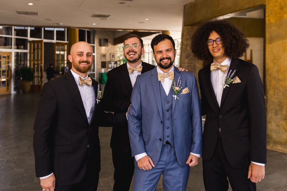 Four people in formal attire pose together in a hallway, wearing suits in blue and black with boutonnieres.