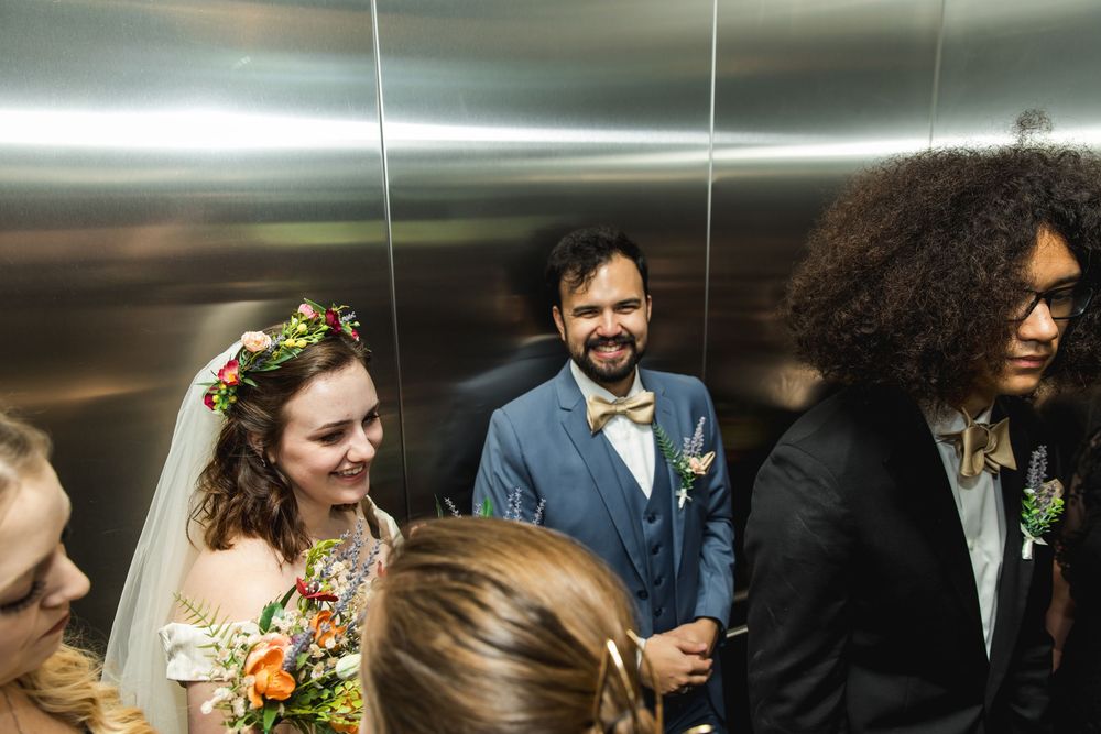 Wedding party members stand close together and smile while riding in a crowded elevator with floral decorations.