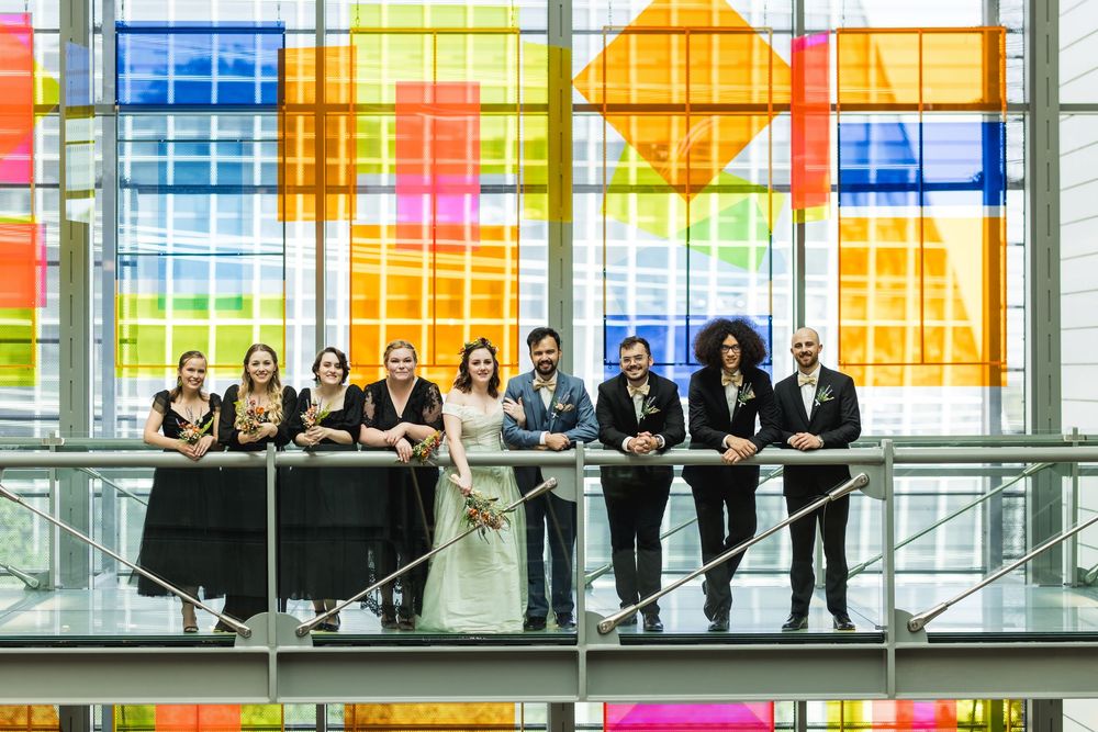 Wedding party stands against vibrant multicolored glass windows in modern building atrium.