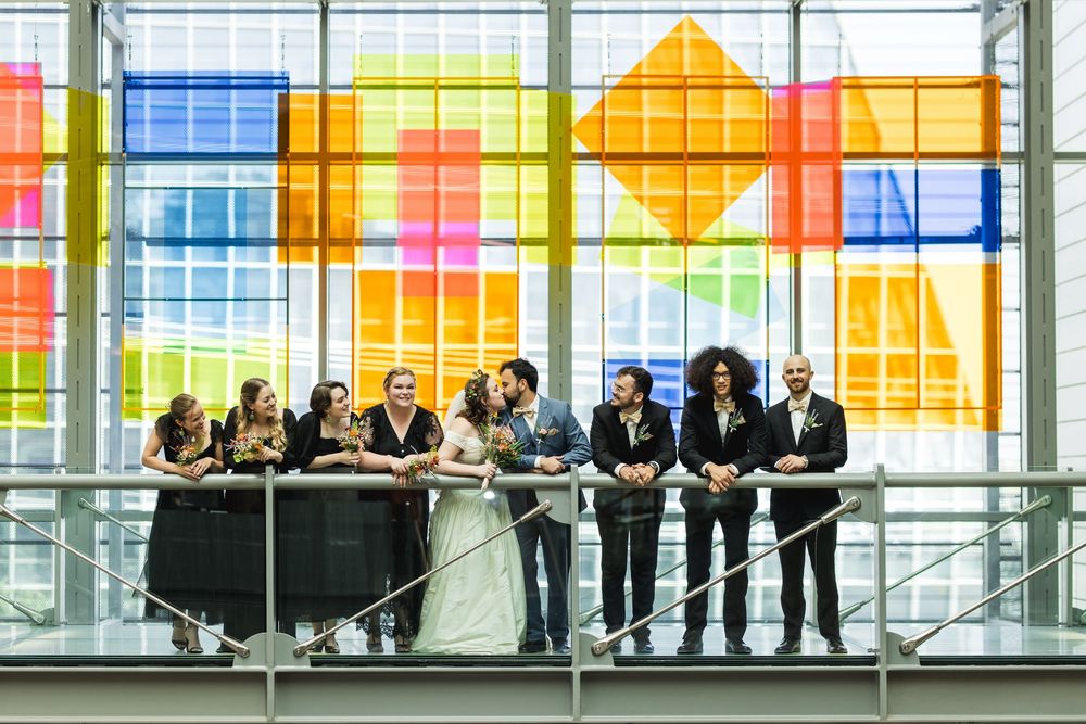 Wedding party poses on a balcony against colorful geometric stained glass windows in modern architectural setting.