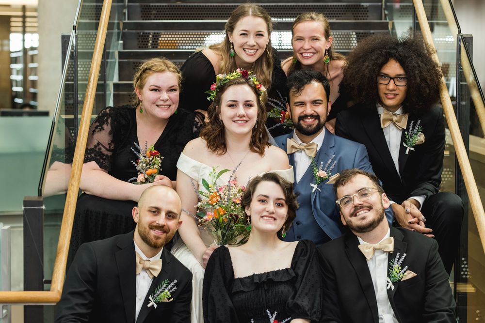 Wedding party members in black and navy attire pose together on stairs with bouquets and boutonnieres.