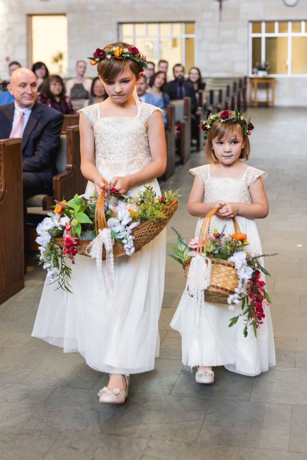 Children in white flower girl dresses walking down a church aisle holding small floral bouquets during a wedding ceremony.