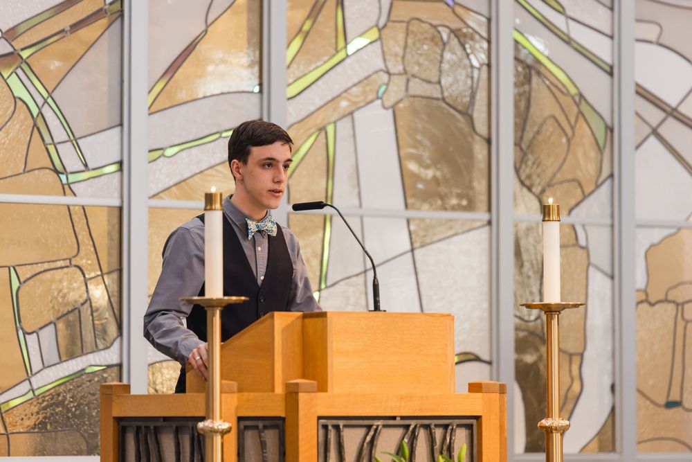 A speaker stands at a wooden podium in front of an abstract stained glass window backdrop.