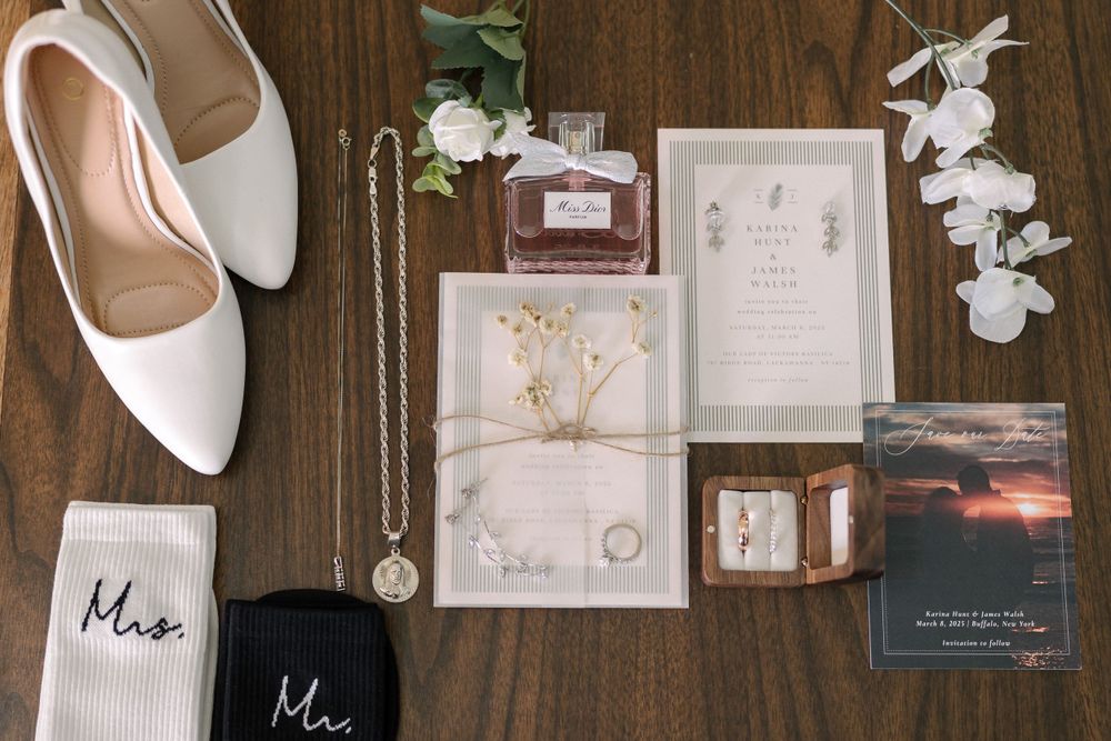 Wedding details flatlay with white shoes, floral invitation suite, Mr and Mrs socks, and perfume on wooden surface.