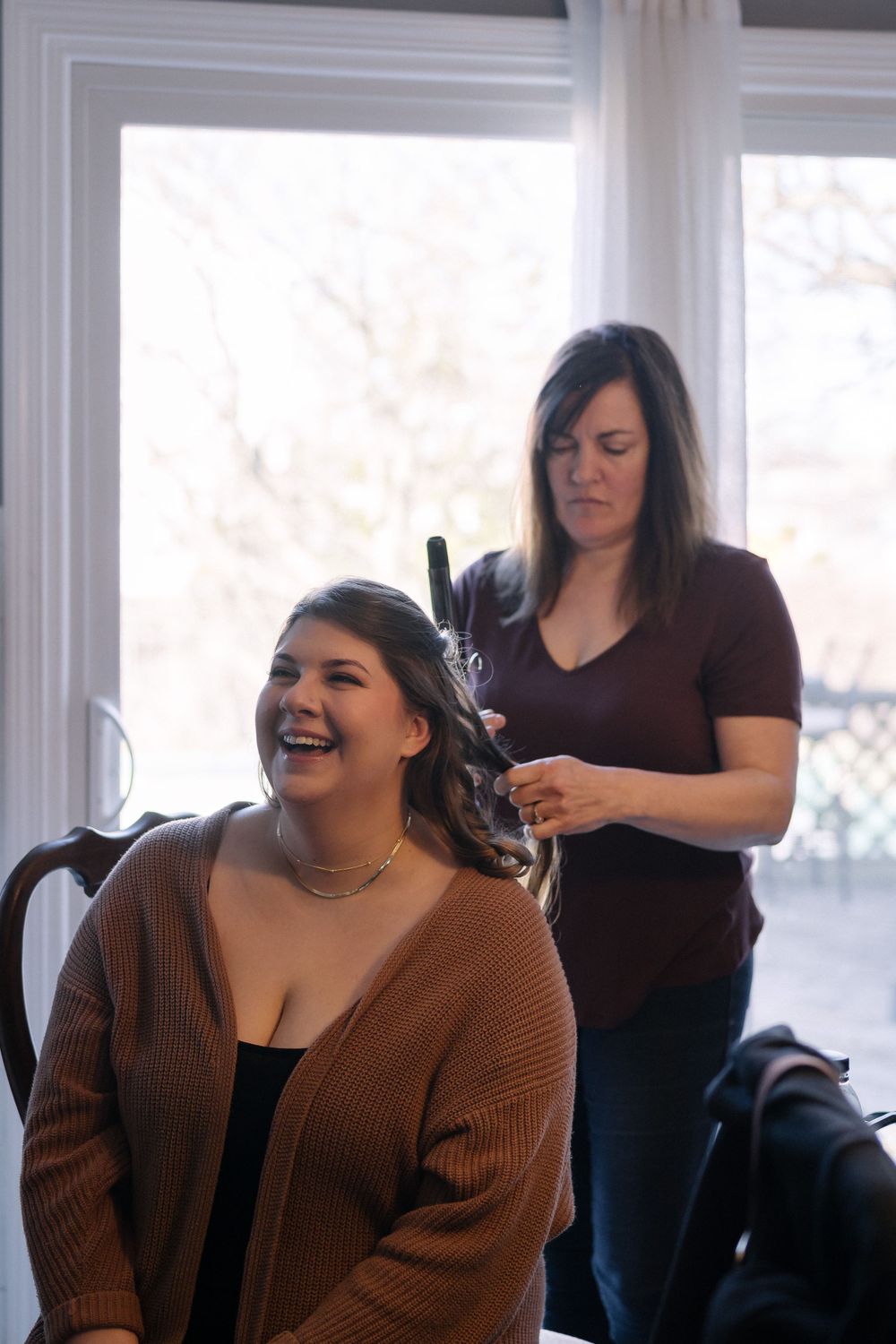 A hairstylist styles a client's hair by a bright window while both share a joyful moment together.