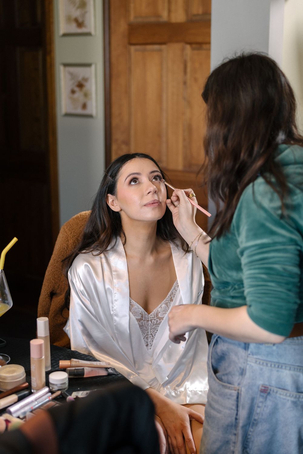 Makeup artist applies eye makeup on a woman in white silk robe sitting at vanity with cosmetics spread out.
