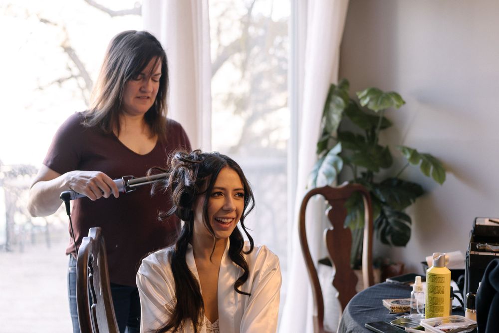 A stylist works on a client's hair near a bright window with a houseplant in the corner.