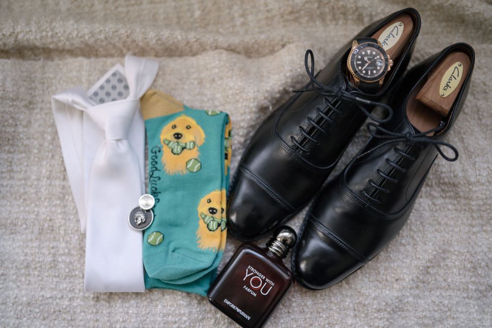 Black leather dress shoes arranged with white socks, colorful patterned socks, and a small bottle of fragrance on textile backdrop.