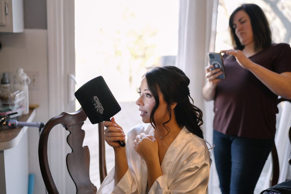 Someone gets ready in the morning using a handheld mirror while sitting at a table while another person checks their phone.