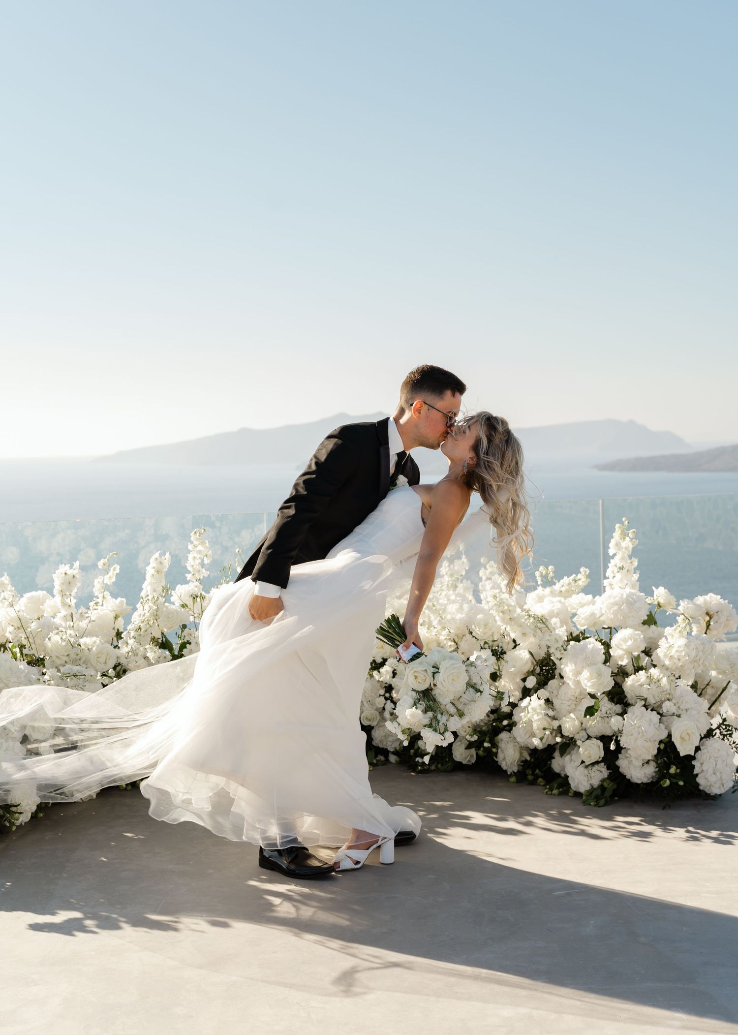 Romantic wedding couple shares a kiss against stunning ocean view backdrop with elegant white floral arrangements.