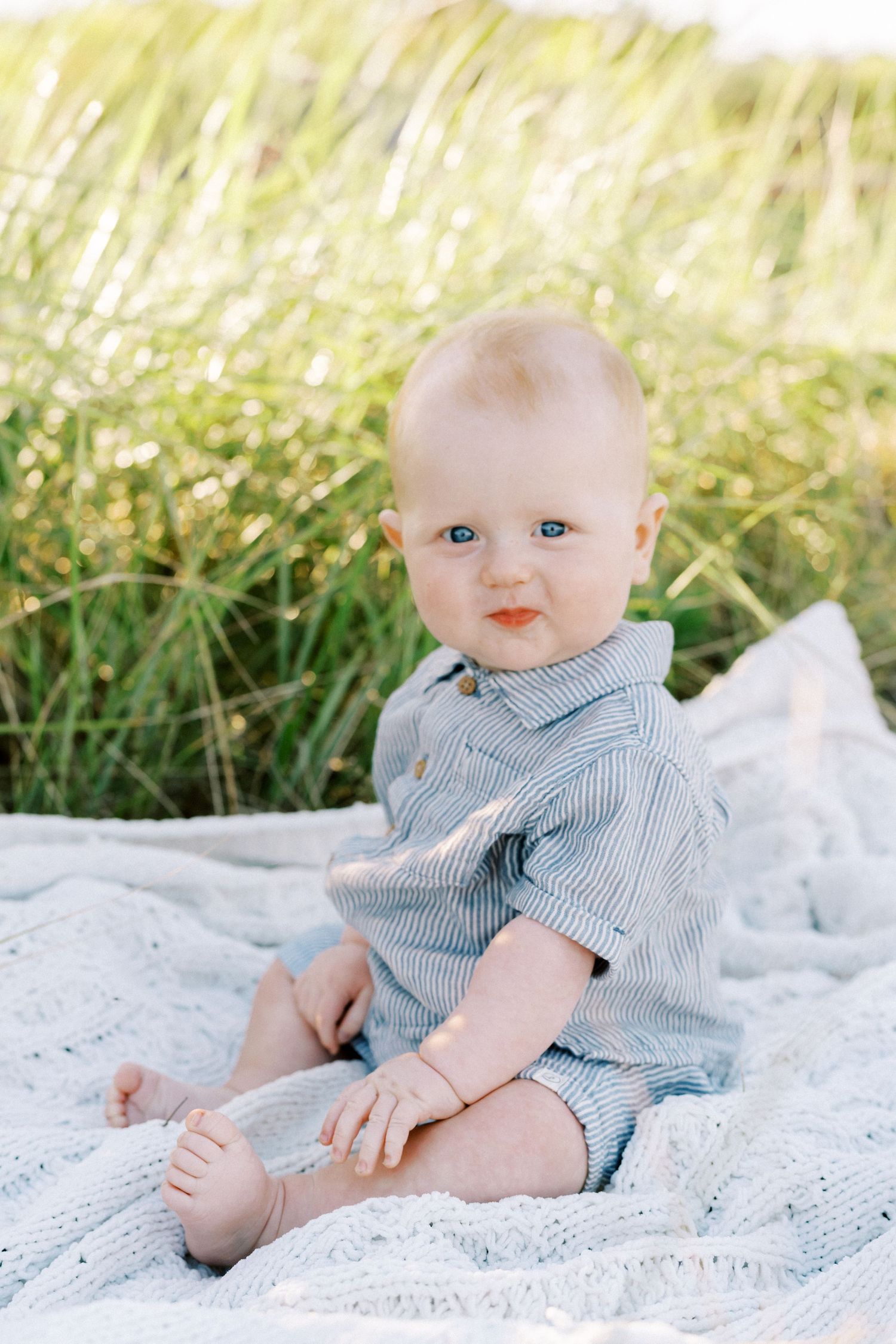 Baby in blue striped romper sitting on white blanket with tall grass in background.
