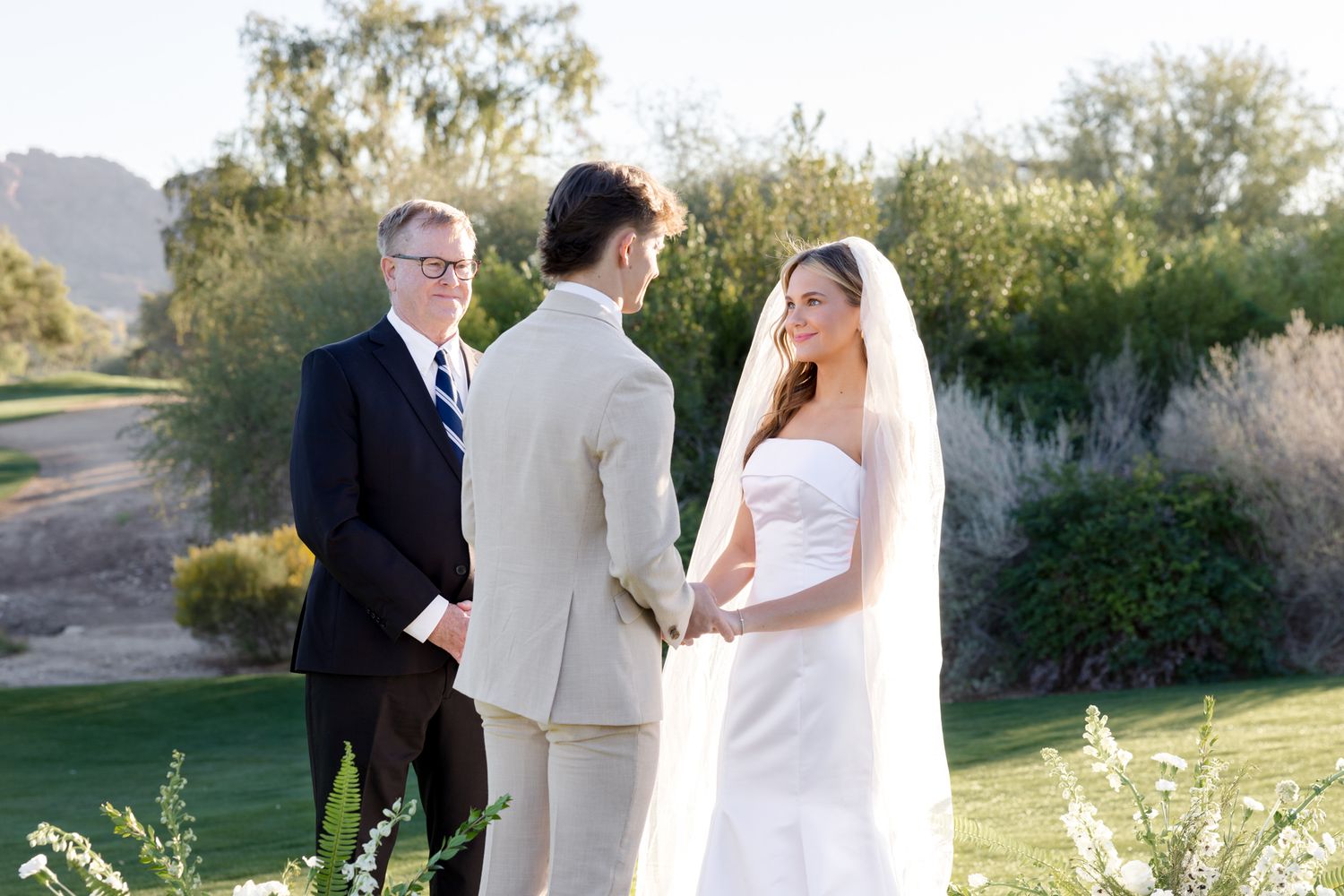 A wedding ceremony takes place outdoors on a scenic golf course with desert mountains and greenery in the background.