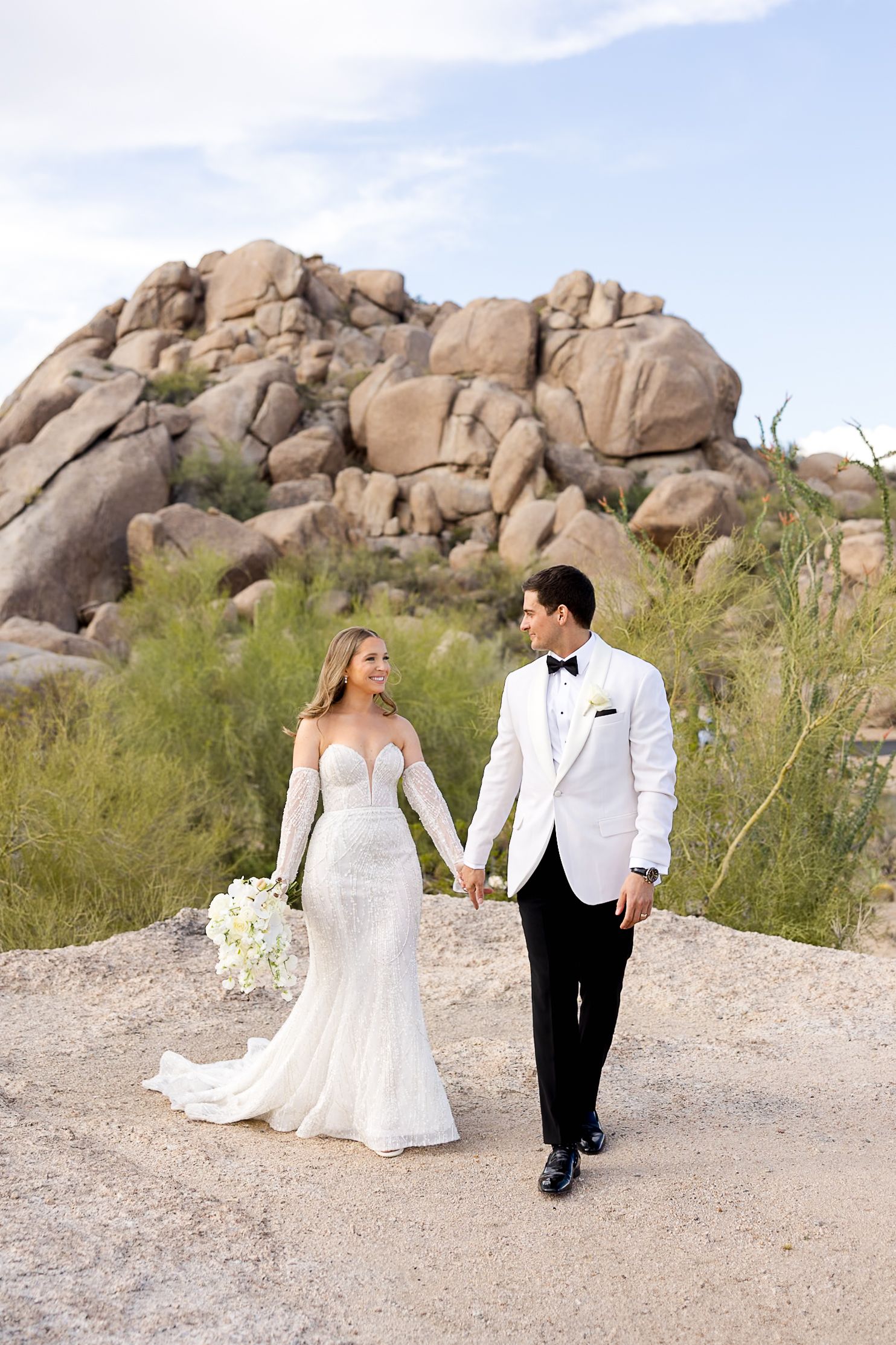 A bride and groom walk and pose together on desert terrain with large boulder formations in the background.