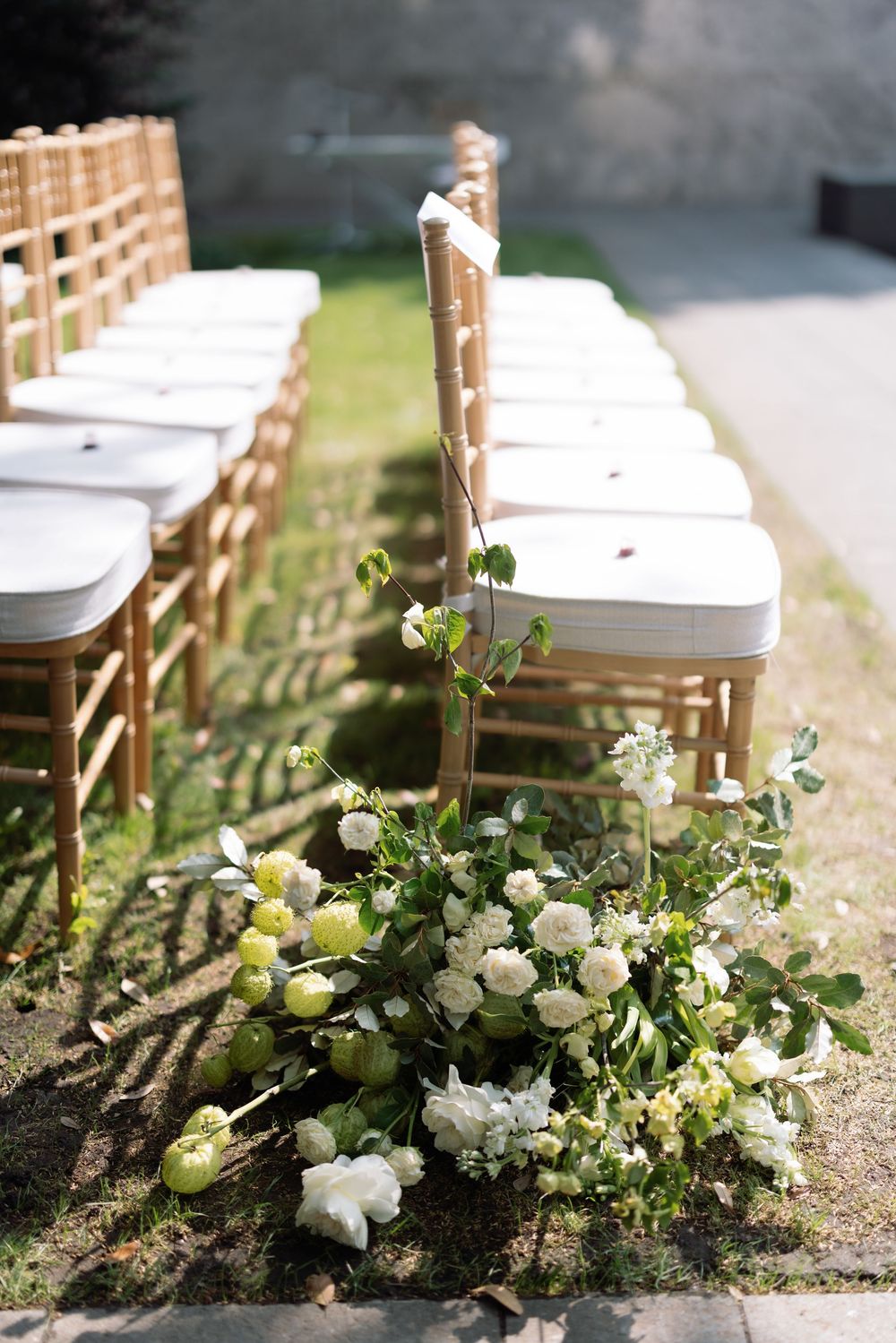A series of white floral wedding aisle decorations with greenery arranged along white folding chairs at an outdoor ceremony.