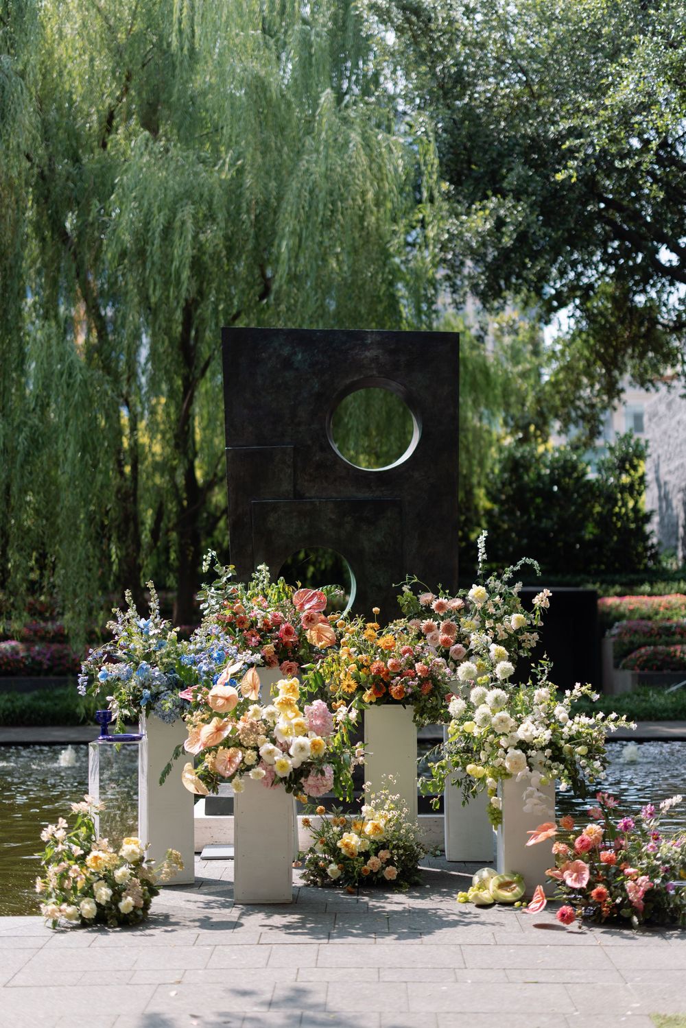 Elegant outdoor wedding ceremony setup with white pedestals holding colorful floral arrangements near a willow tree and pond.