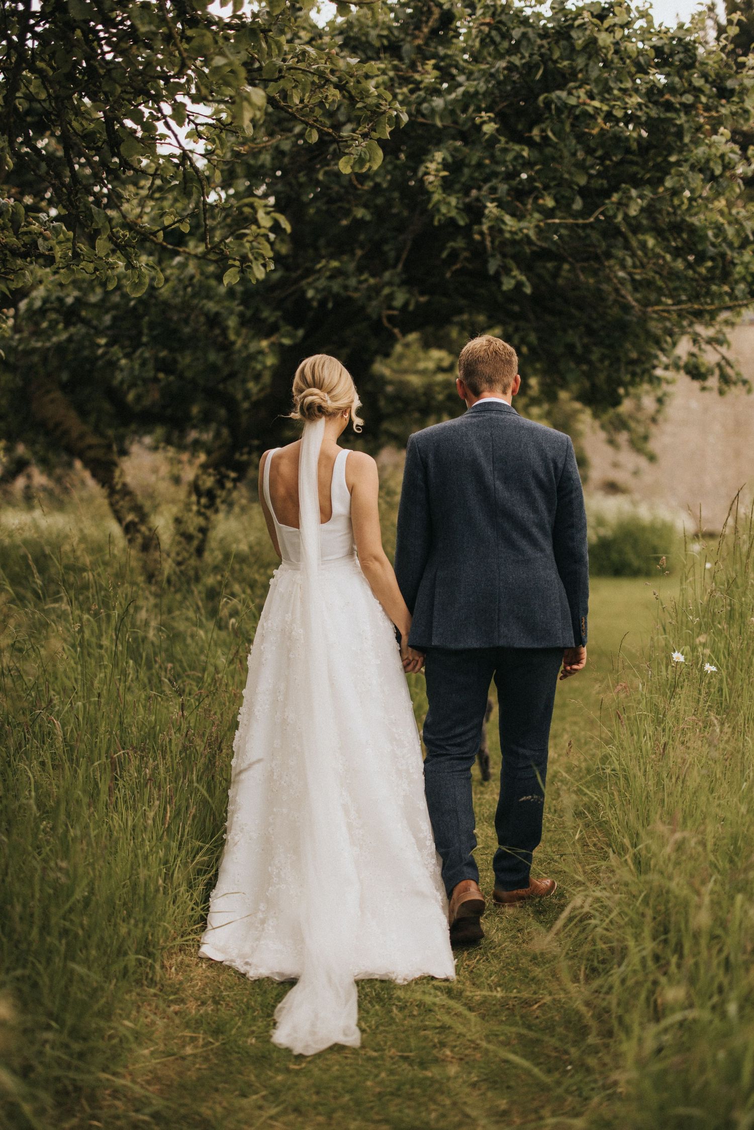 Newlywed couple walking through tall grass under a tree, with bride in flowing white dress and groom in suit at Scrivelsby Walled Garden.