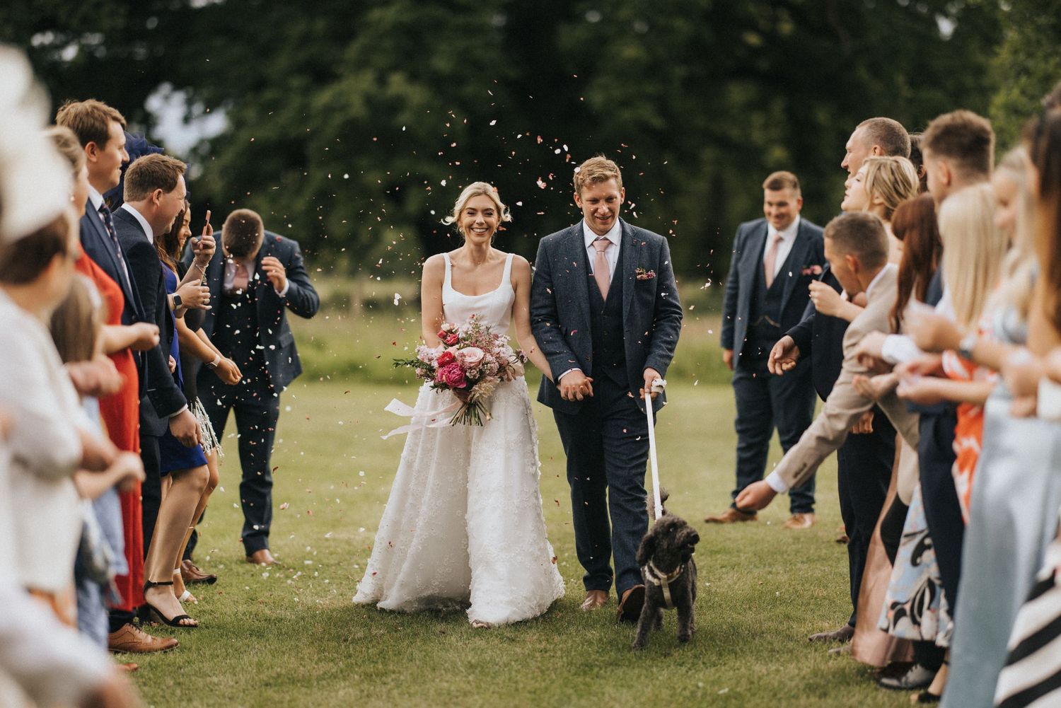 Couple walks down grassy aisle with their black dog while wedding guests toss confetti in celebration outdoors at Scrivelsby Walled Garden.