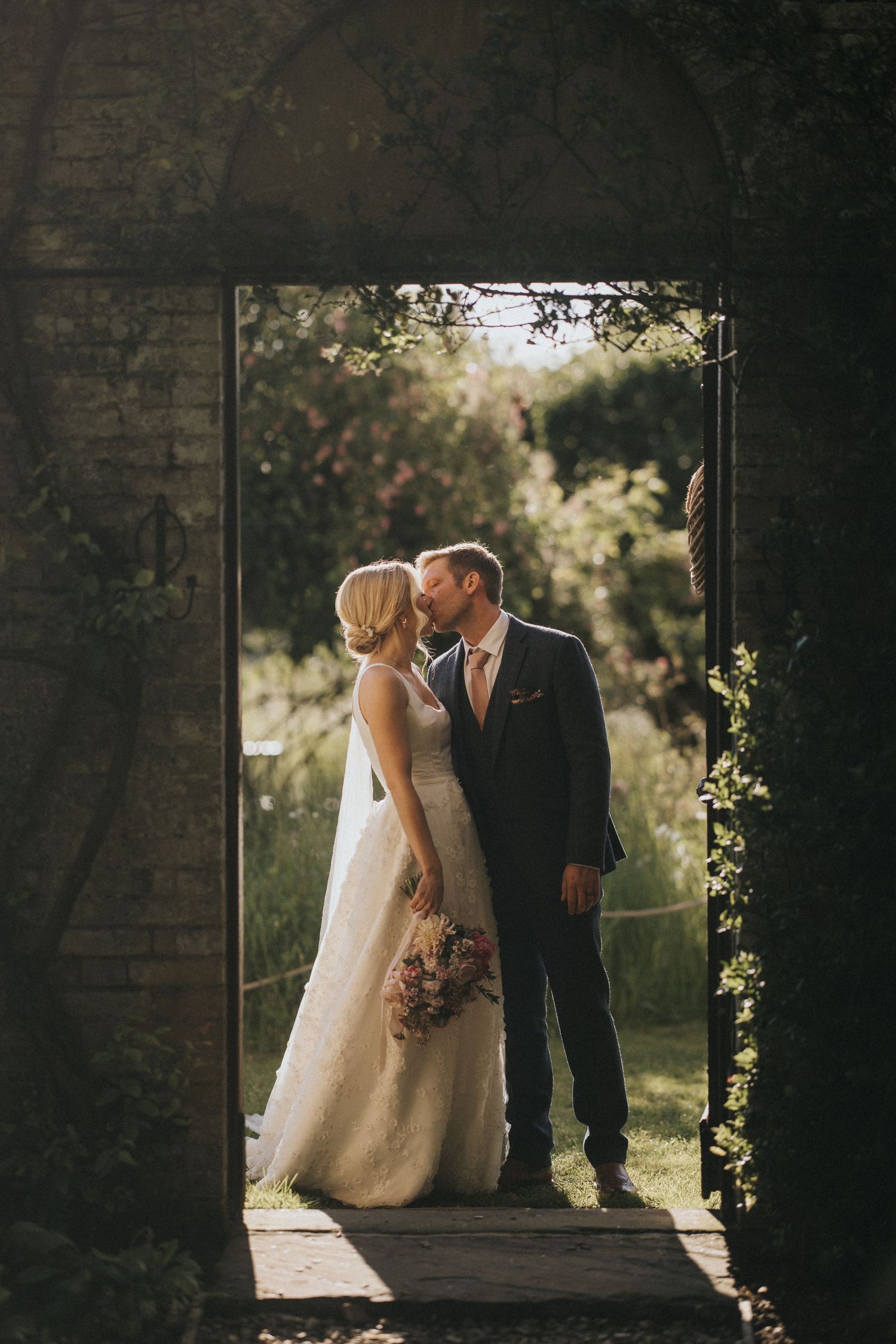 Romantic wedding couple embrace in sunlit stone doorway surrounded by natural greenery and backlit glow at Scrivelsby Walled Garden.