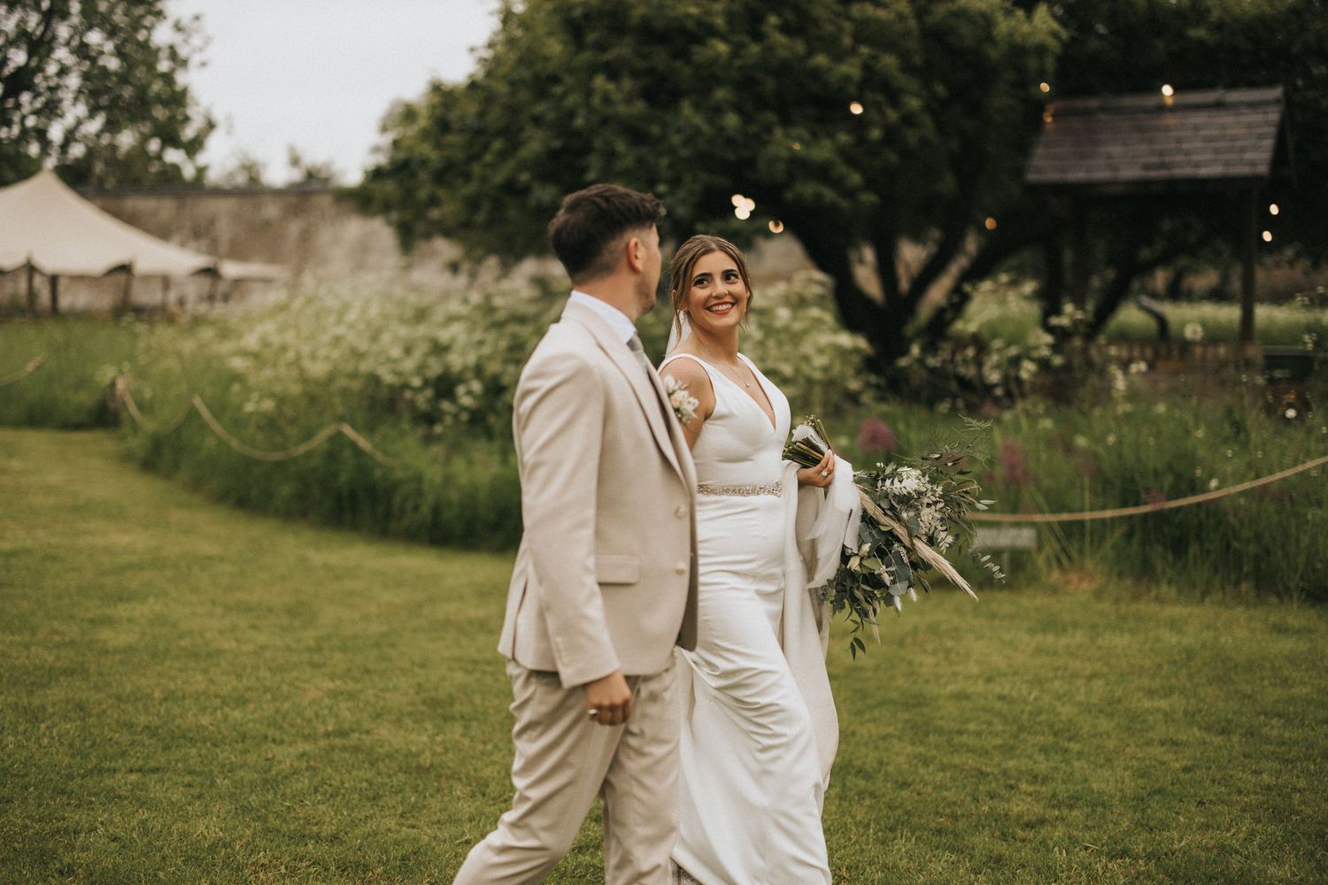 Newlywed couple in light beige and white attire walking together on green lawn during wedding celebration at Scrivelsby Walled Garden.