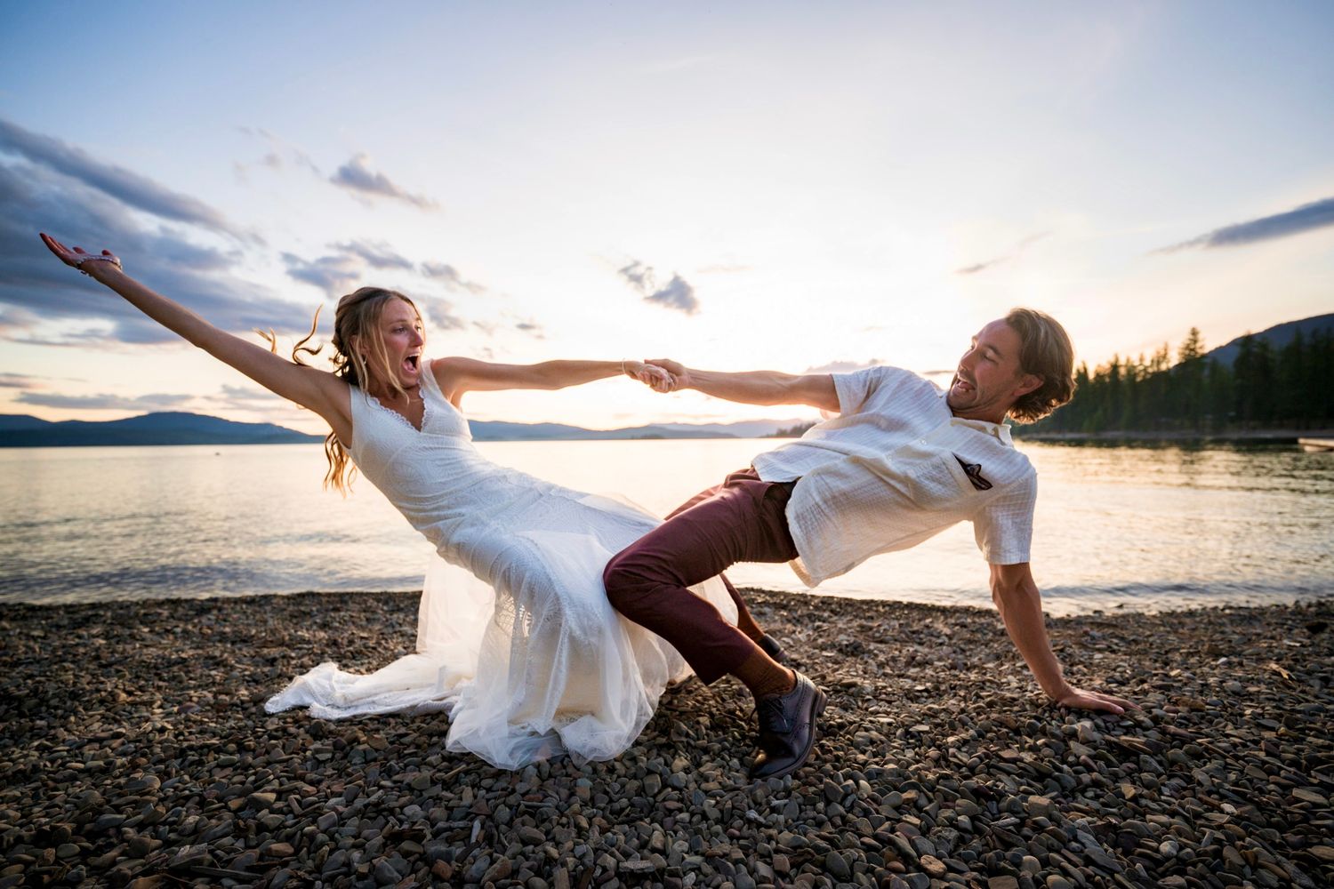 Playful couple dances together on a scenic beach at sunset with mountains and water in the background.