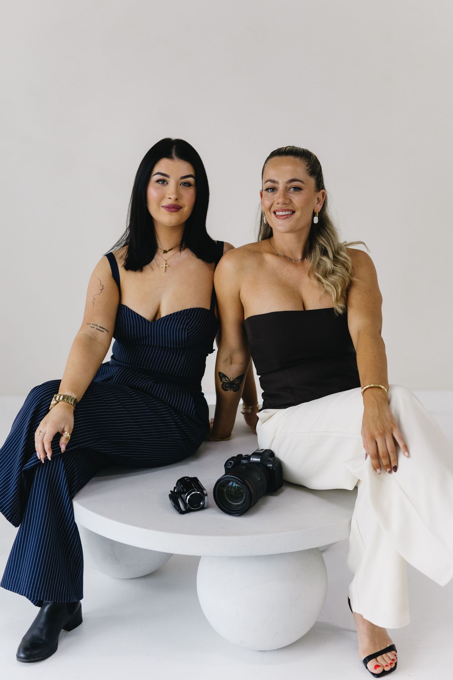 Two photographers sit on a modern white table with DSLR cameras wearing contrasting black and white outfits.