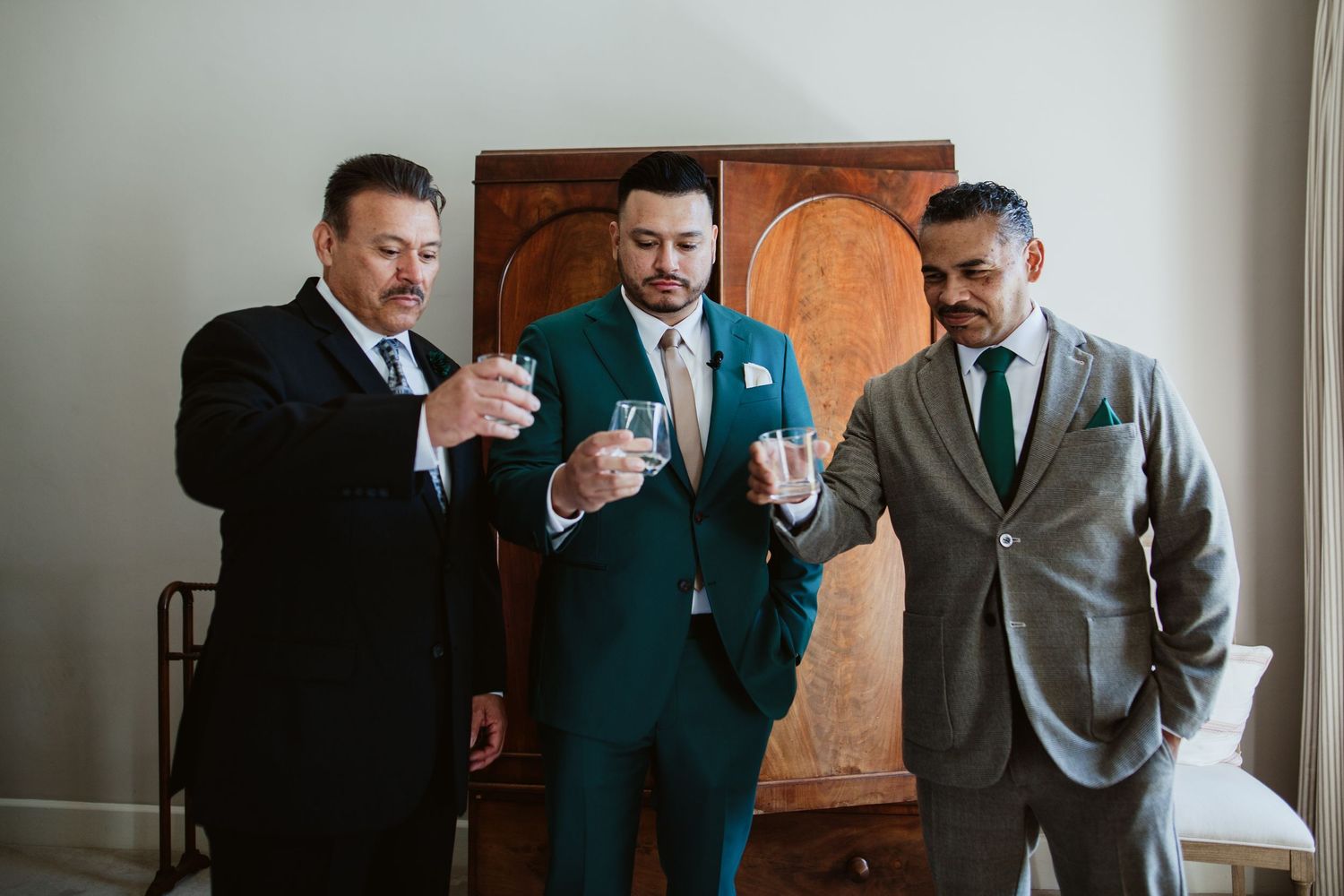 Three men in formal suits look at a smartphone during a wedding preparation, standing in front of a wooden armoire.