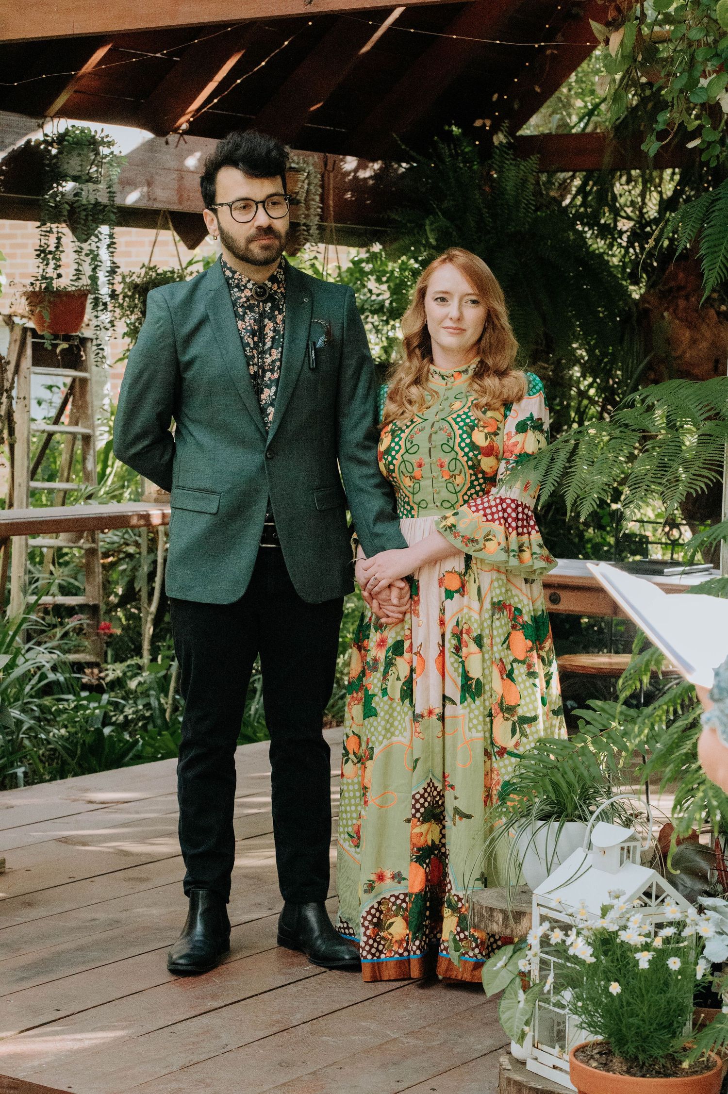 A couple poses on an outdoor wooden deck dressed in green and floral attire among garden decorations and lanterns.