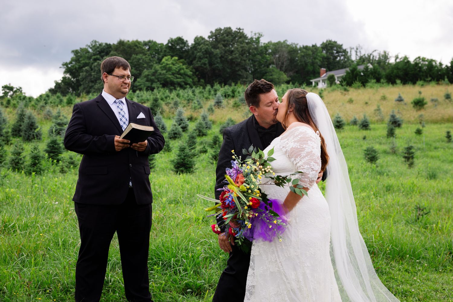 An outdoor wedding ceremony takes place in a green field with a couple and officiant against rolling hills and trees. Tennessee Tree Farm.