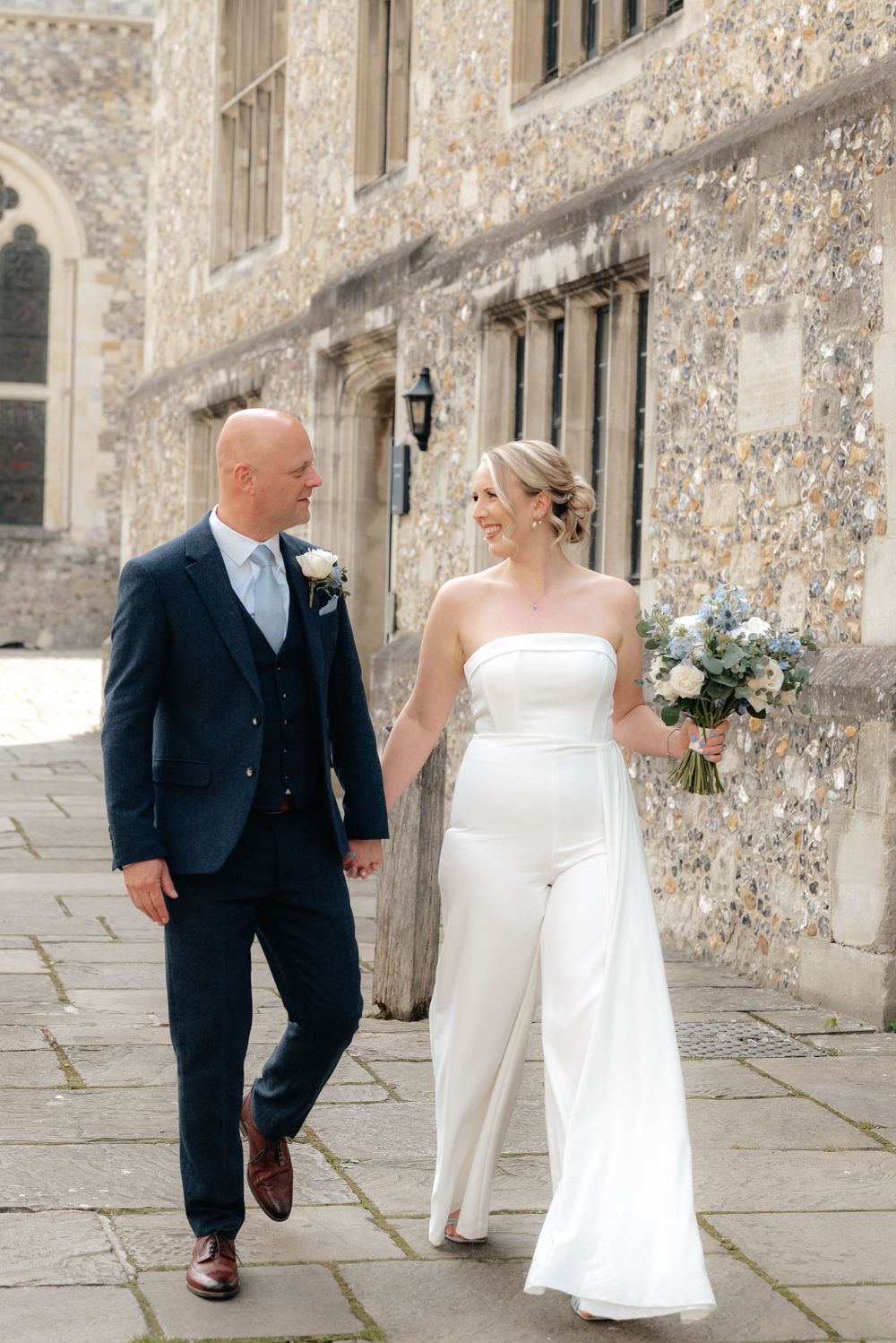 Couple walking together along stone building exterior during wedding celebration.