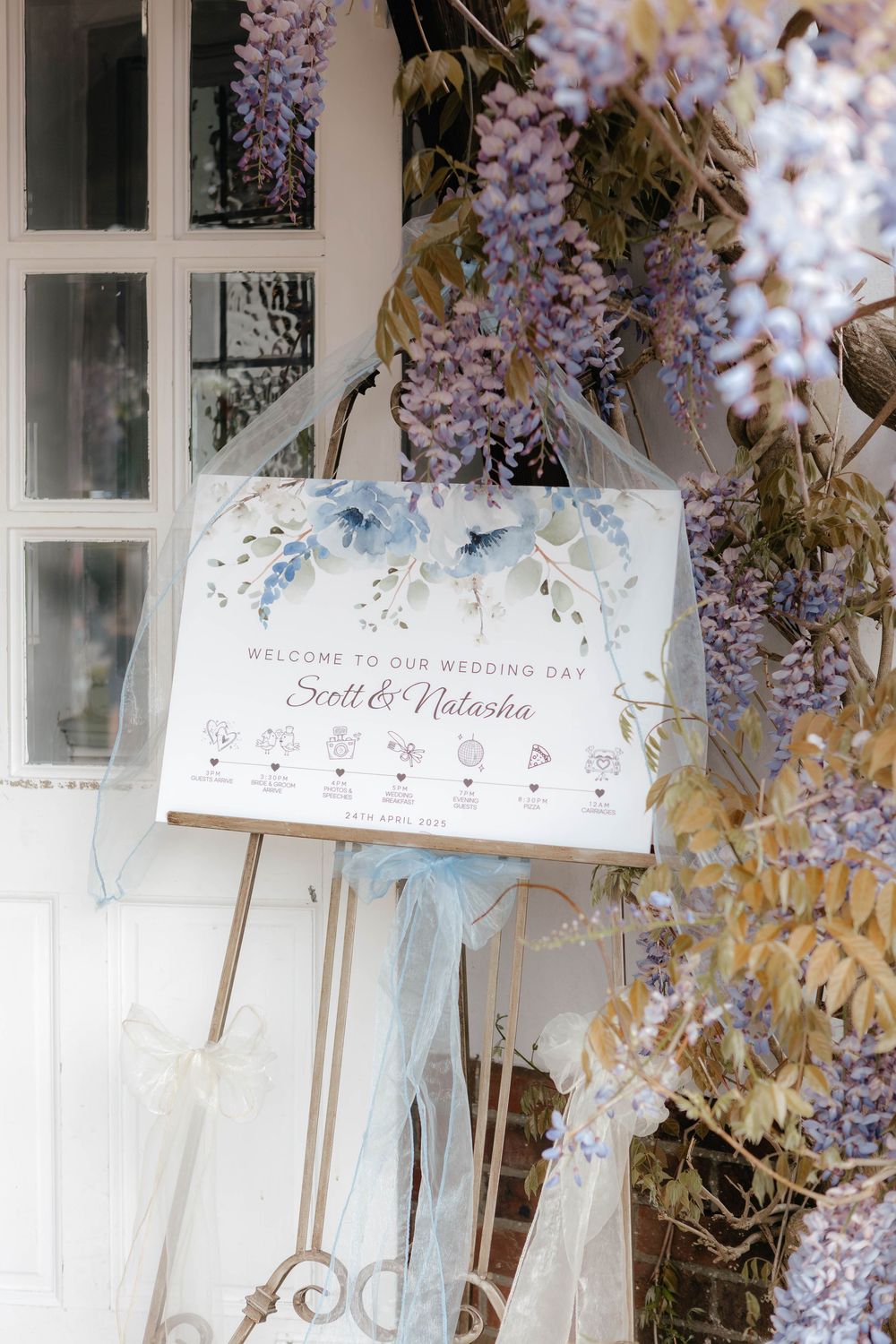 White decorative signs with script text hang from purple wisteria vines against a light-colored wall.