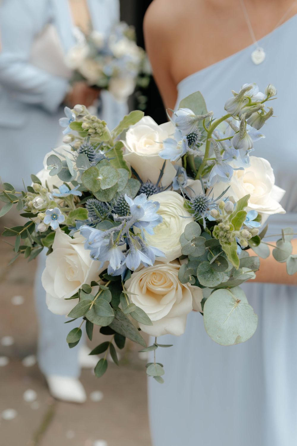 Elegant wedding bouquet with white roses, blue thistles, and green eucalyptus leaves in a soft pastel color palette.