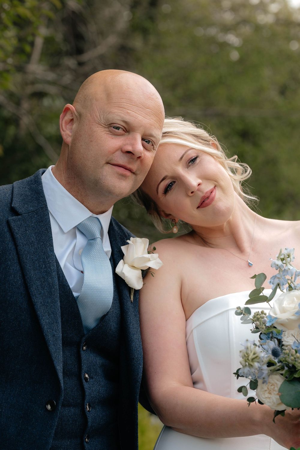 Portrait of a happy couple on their wedding day wearing formal attire and holding a blue and white floral bouquet.
