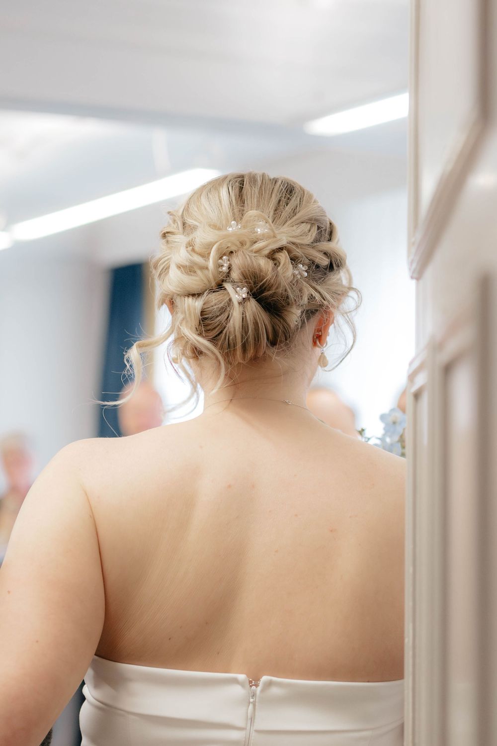 An elegant updo hairstyle with soft blonde curls and delicate pearl details, shown from behind in a white strapless dress.