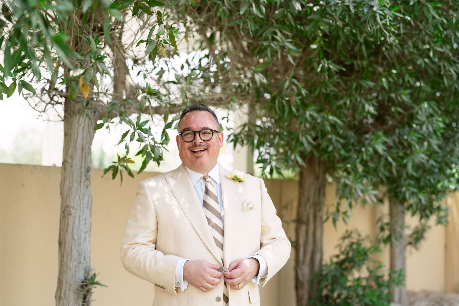 A person in a light suit and tie stands against a beige wall with green foliage visible nearby.