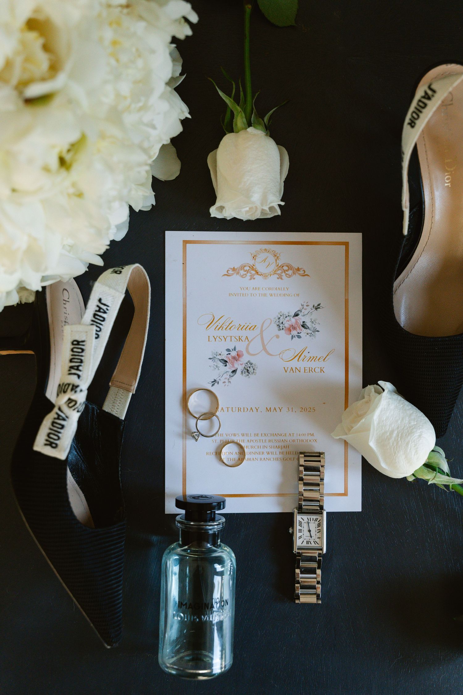 Wedding invitation with floral design displayed alongside wedding rings, white roses, a blue mason jar, and shoes on black surface.