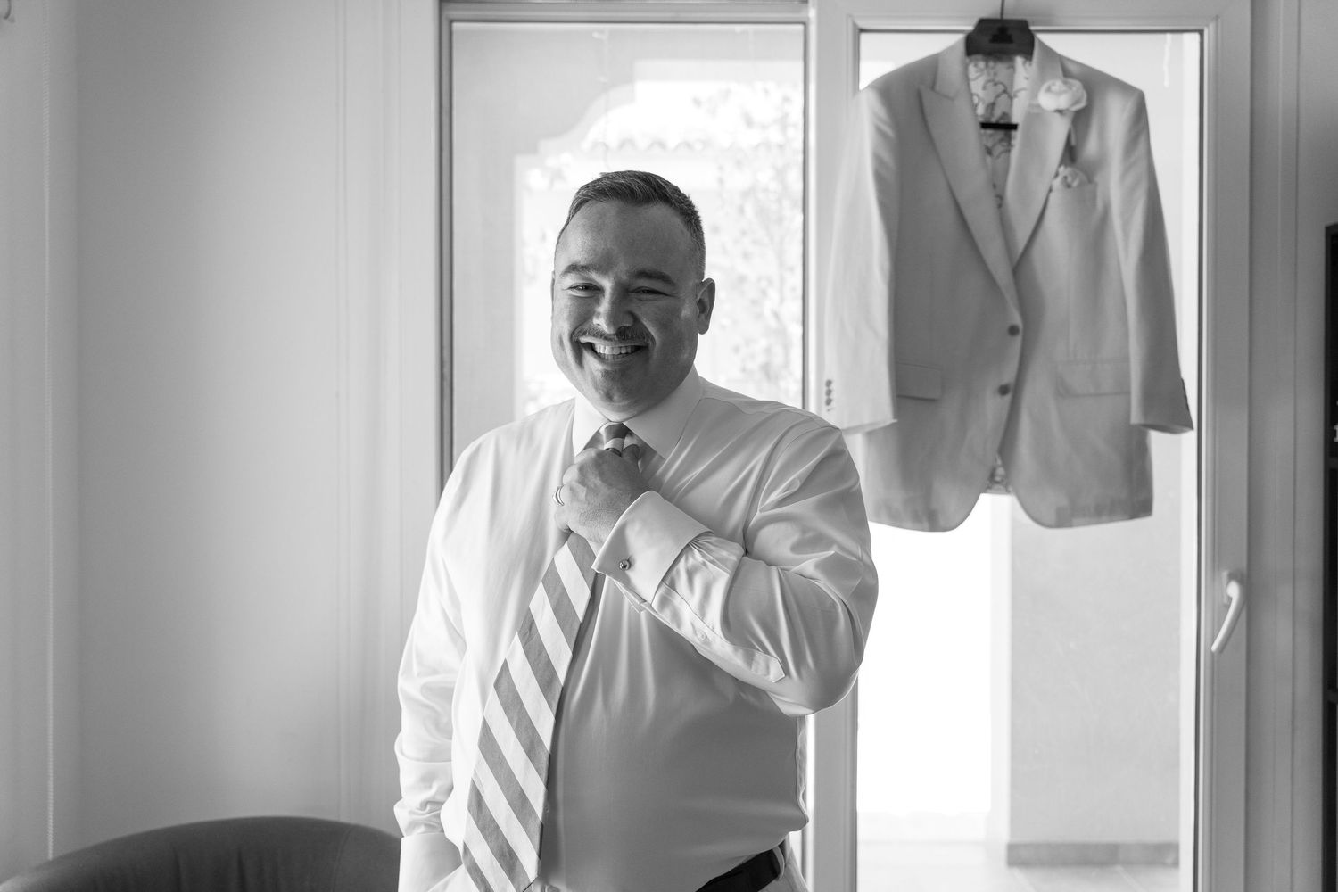 Black and white photo showing someone adjusting their tie while standing in a room with a suit jacket hanging on the wall.