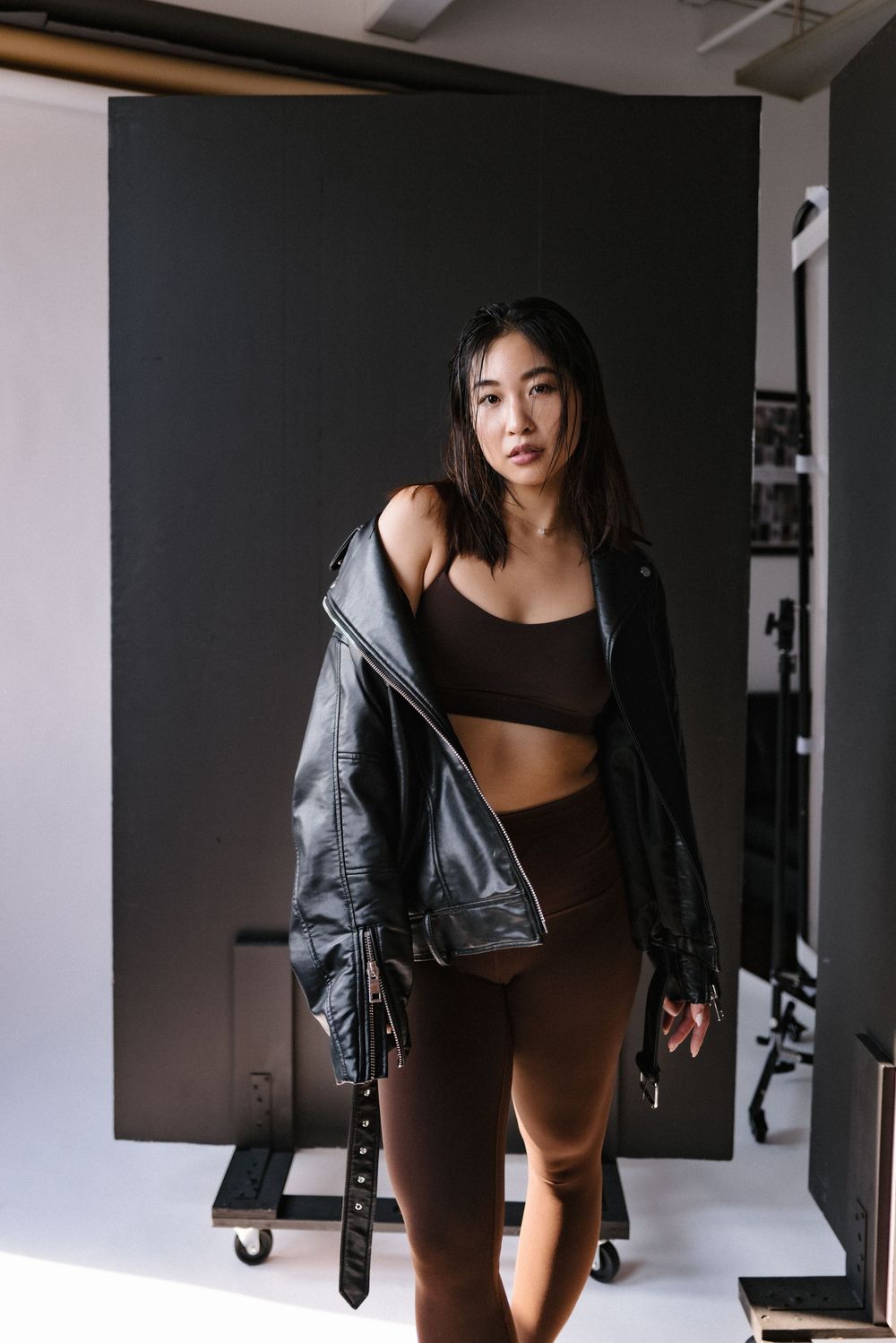 Athletic fashion shoot featuring dark leather jacket over sports bra and brown leggings in a minimalist studio setup.