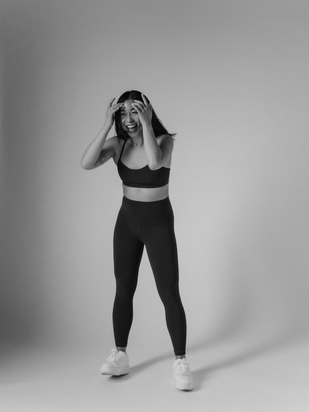 A black and white photo of a person in athletic wear and white sneakers posing with hands covering their head.