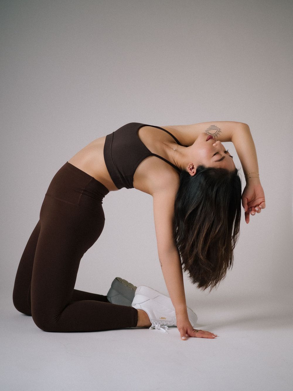 Athletic figure in dark workout attire demonstrates a yoga camel pose against a light gray studio background.