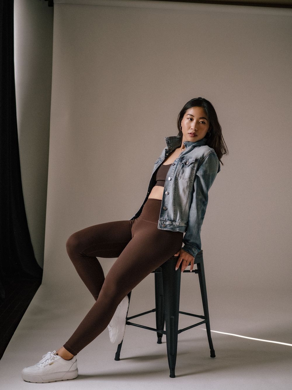 Athletic model in denim jacket and brown leggings poses on bar stool against neutral studio backdrop.