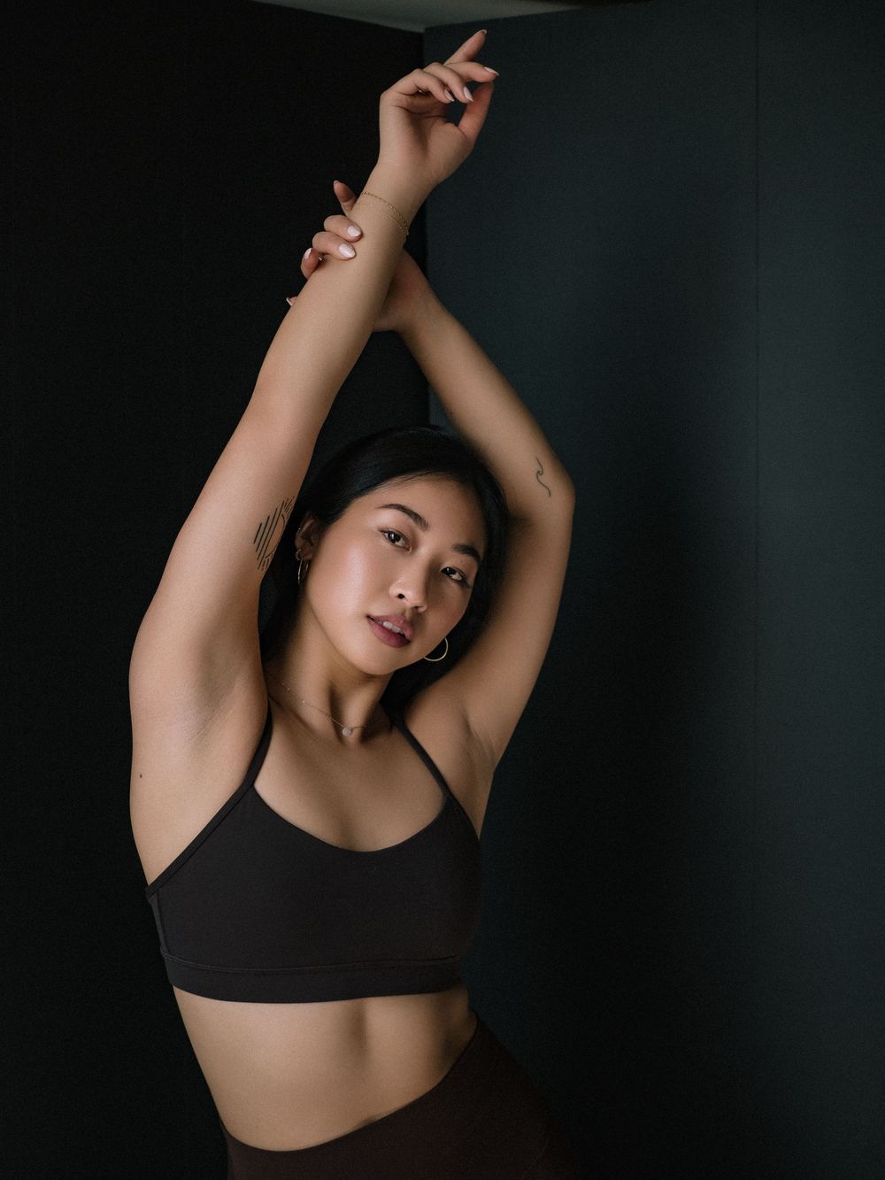 Athletic woman in black sports bra stretches with raised arms against dark backdrop in moody portrait.