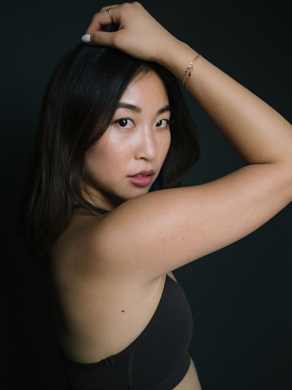 Portrait against a dark background of a person with long black hair wearing a black top and delicate bracelet.